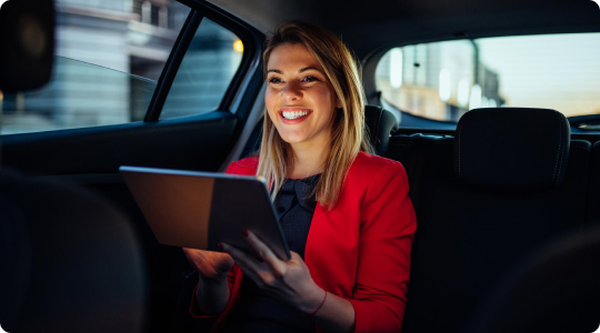 Smiling woman in a red blazer using a digital tablet in the backseat of a moving car during evening hours.