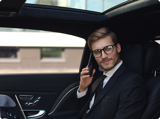 Smiling executive in a suit on a phone call, seated comfortably in the rear of a luxury vehicle with sunroof and leather interior.