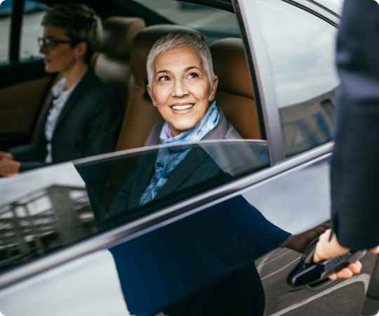 Professional woman smiling warmly as she looks out from the back seat of a chauffeured car while the driver opens the door.