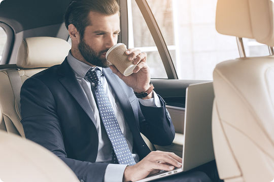 Executive man in a suit working on a laptop and sipping coffee in the backseat of a car with beige interior.