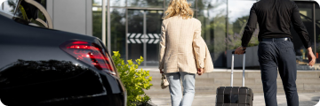 Man and woman walking away from a black luxury vehicle, pulling rolling luggage toward a building entrance