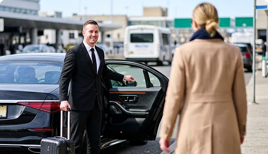 A smiling chauffeur in a suit holds open the door of a luxury car for a woman in a green dress walking toward him. The shot is taken from inside the vehicle looking out onto the passenger pickup curb outside an airport terminal building.