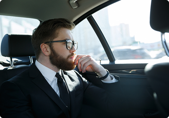 A bearded man in a black suit and tie gazes out the city window from a black car's backseat, deep in thought and sunlight.