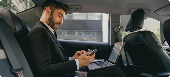 Young businessman multitasking in the backseat of a car, checking his phone while working on a laptop with a coffee cup nearby.