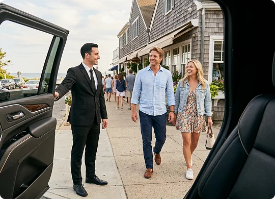 A smiling chauffeur in a suit holds open the door of a luxury vehicle to greet an approaching couple. The perspective from inside the car shows a man in a light blue shirt and a woman in a floral dress walking on a seaside town sidewalk.
