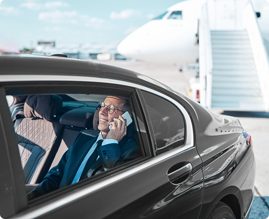 Senior executive seated in a luxury car near a private jet, smiling while talking on the phone, with airplane stairs visible in the background.