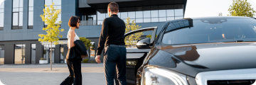 Chauffeur greeting a woman beside a luxury car outside a commercial building.