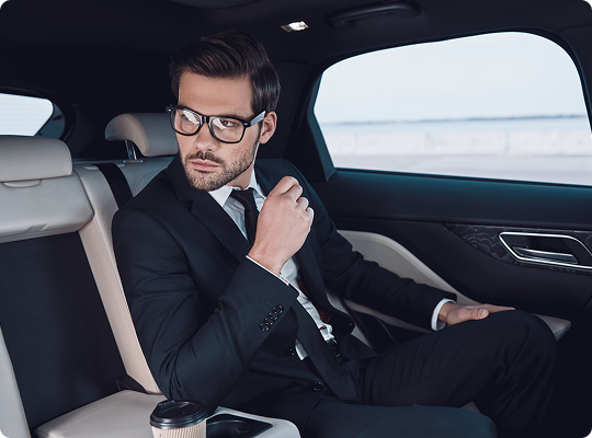 Stylish executive with glasses seated in the rear of a luxury car, gazing out the window with a coffee cup beside him.