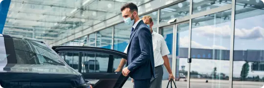 A business travelers in masks step into a sleek black car outside a modern airport terminal, capturing safe, professional airport transfers.