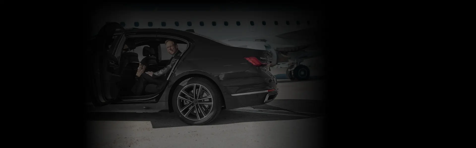 Rear-angle view of a luxury sedan parked near a plane with a smiling passenger, highlighting seamless EWR Airport Transfers in style.