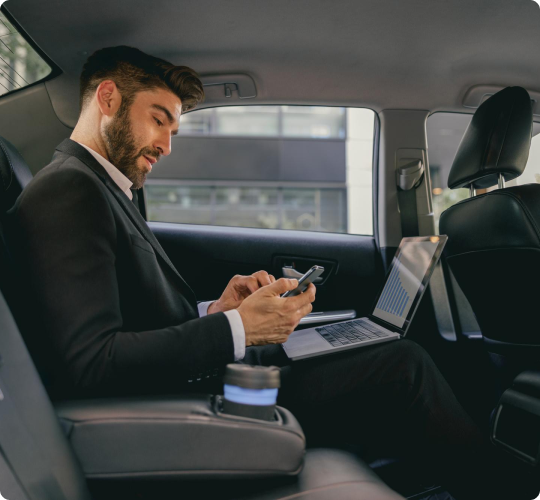 Businessman in a black suit using a smartphone with a laptop open beside him and a coffee cup in the armrest, working while riding in the back of a car.