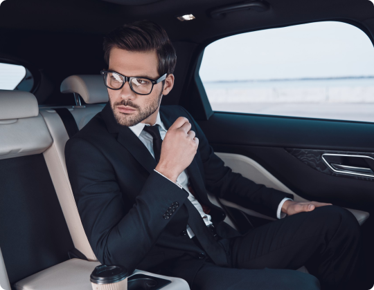 Stylish businessman in a suit and glasses seated in the back of a luxury car, gazing out the window with a coffee cup beside him