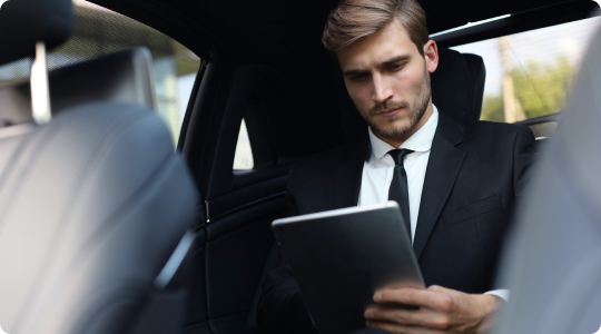 Young professional man in a black suit focused on a tablet while seated in the backseat of a luxury vehicle.