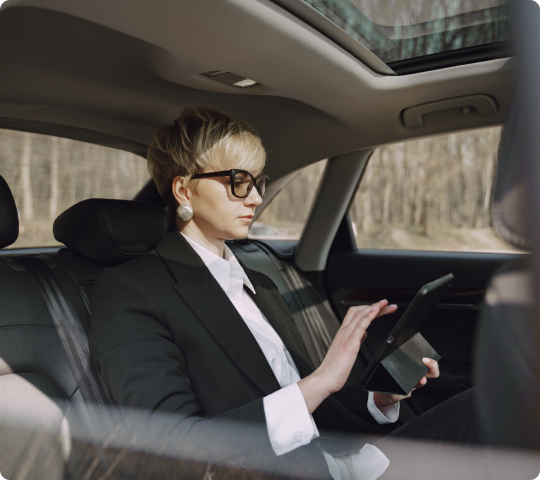 Businesswoman in glasses working on a tablet inside a luxury car, with natural daylight filtering through a sunroof.