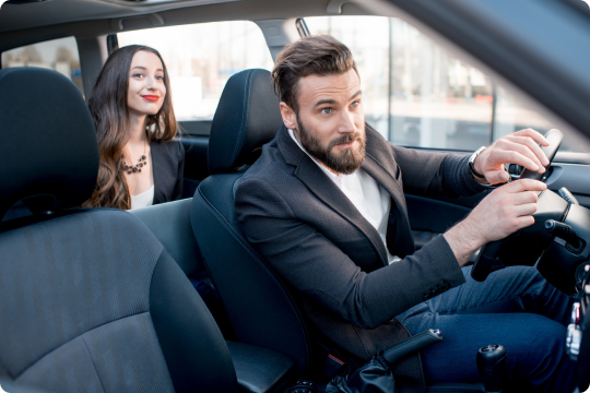 Smiling woman sitting in the backseat of a car, looking at a bearded chauffeur in a suit preparing to drive.