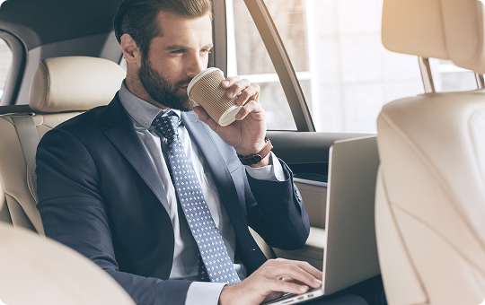 Businessman in a luxury car sips coffee while typing on a laptop, maximizing comfort and productivity on the move.