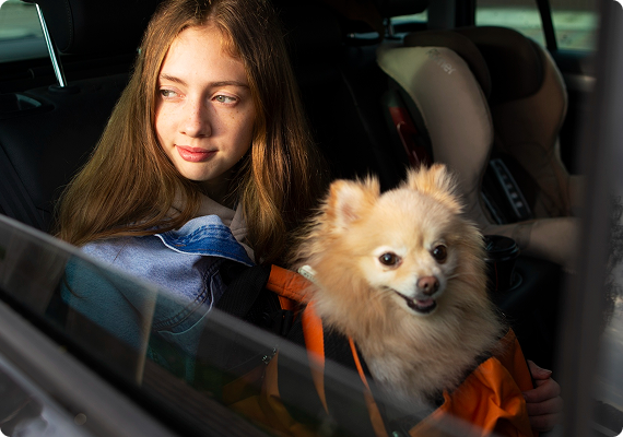 A young girl enjoys a cozy backseat ride with her fluffy dog beside a child safety seat, sunlight streaming through the window.
